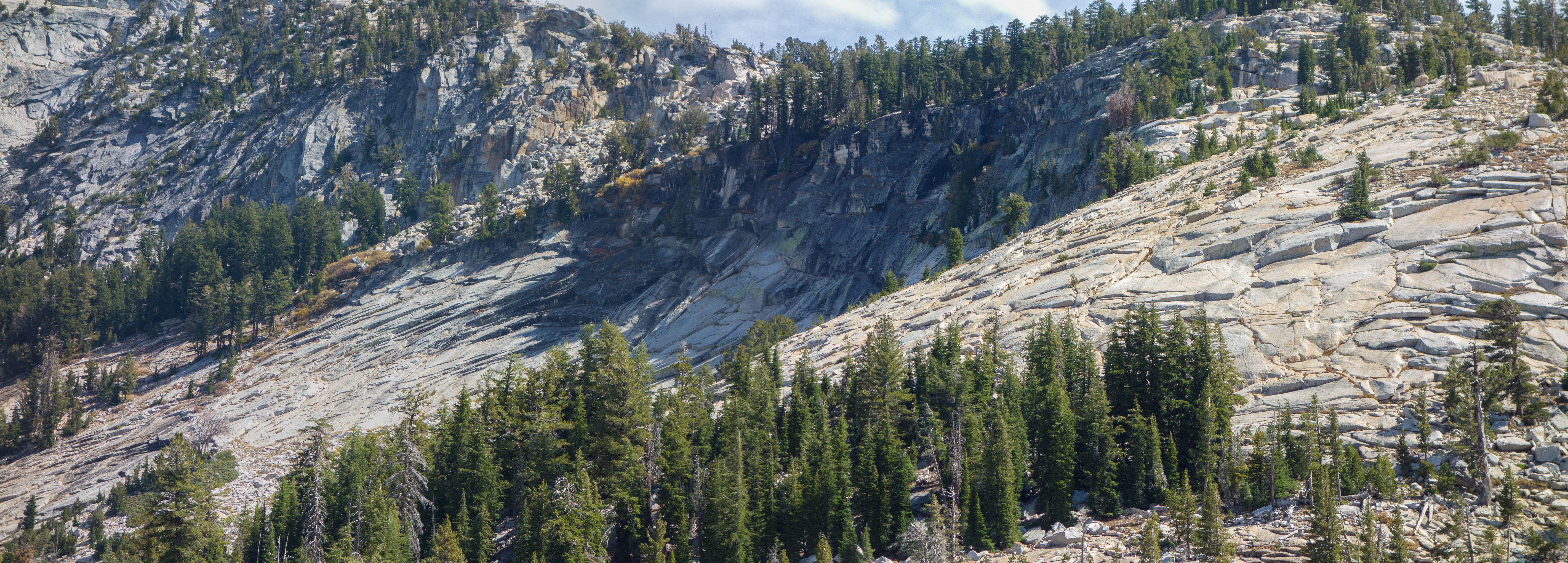 Two possible routes are visible: (1) route enters photo at the left and climbs diagonally upward through the boulder field lying above the cliffs at the center. (2) Route continues across the saddle between the shoulder of Tenaya Peak and the sub-dome, then climbs slabs and continues up the ridge to the summit to the left, out of the photo.